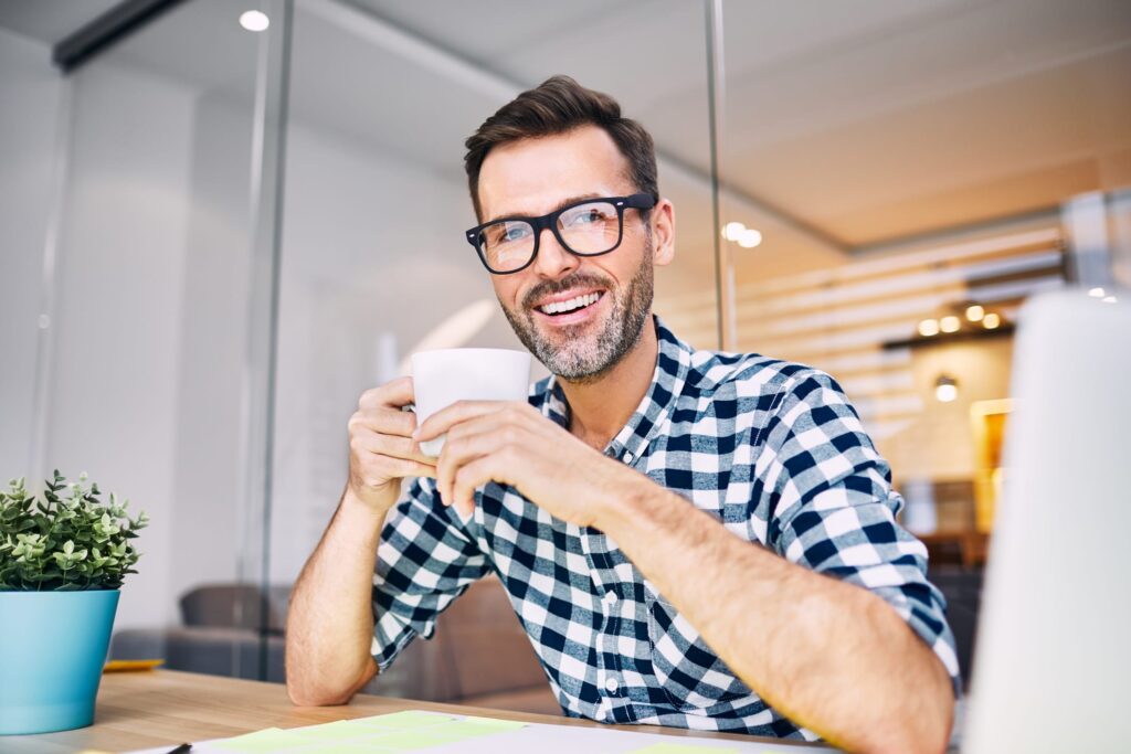 Man sitting at desk smiling and holding a cup of coffee, thinking about relaxing at home with a home massager